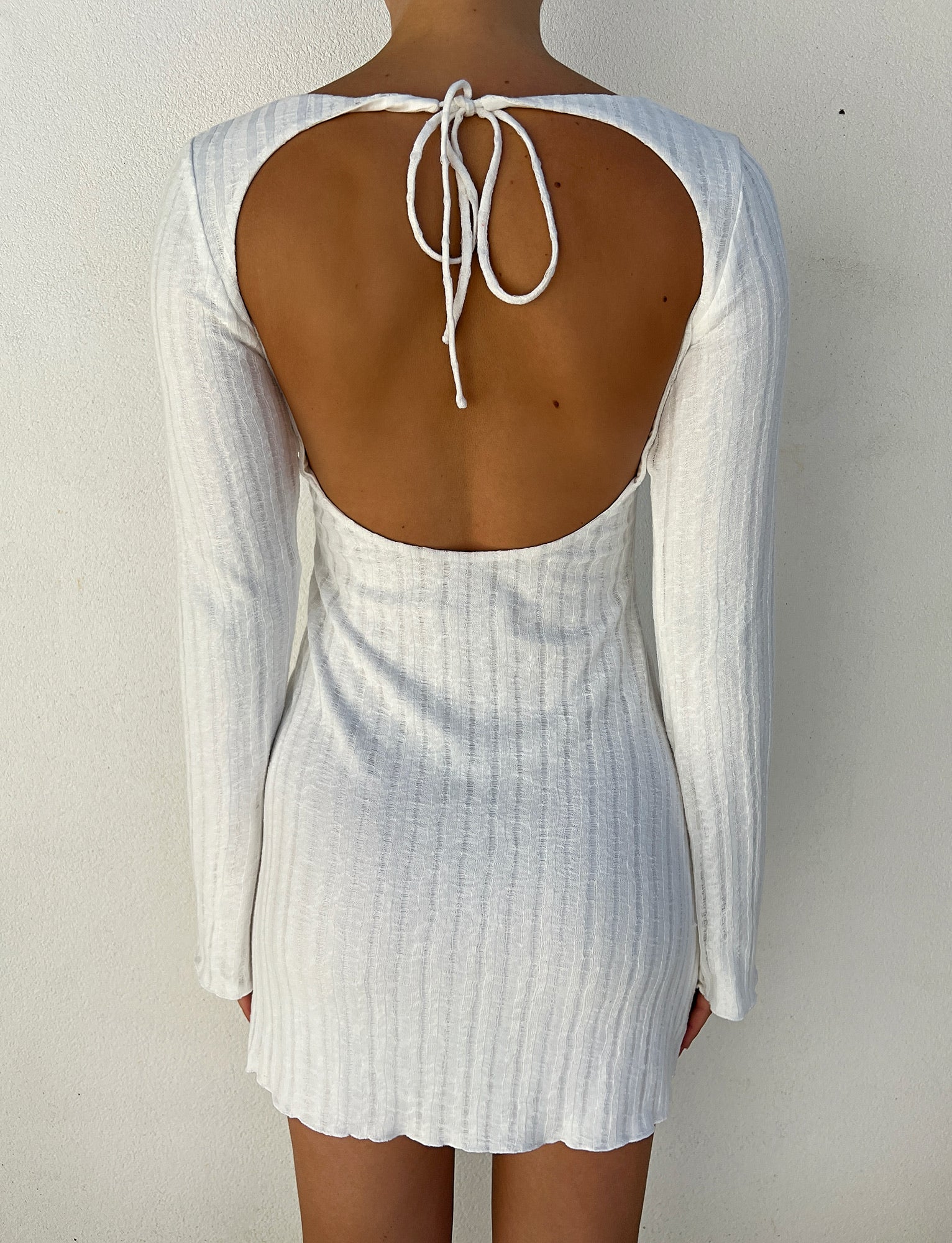 A person is wearing the BRENDEN DRESS - NEUTRAL from TIGERMIST. This long-sleeve, backless white dress features a tie at the neck and a subtle ribbed texture, falling to mid-thigh. The design accentuates the person's fully exposed back against a plain white wall background.