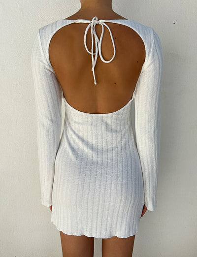 A person is wearing the BRENDEN DRESS - NEUTRAL from TIGERMIST. This long-sleeve, backless white dress features a tie at the neck and a subtle ribbed texture, falling to mid-thigh. The design accentuates the person's fully exposed back against a plain white wall background.