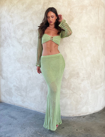 A woman with long brown hair is standing against a textured beige wall, dressed in a light green, crochet knit two-piece outfit from TIGERMIST. The ensemble includes the MACAULEY SKIRT - GREEN: PASTEL GREEN, which features a long-sleeve cropped top and a matching long skirt. She has one hand raised to her head, gazing downward.