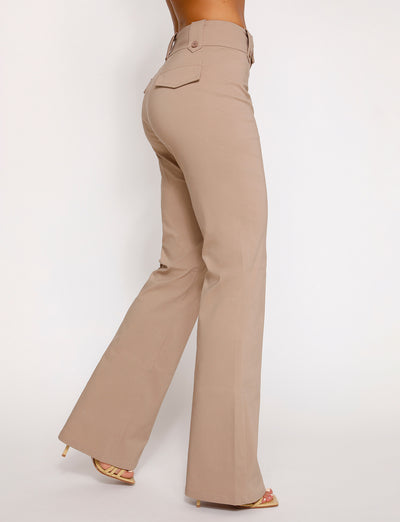 A person is modeling the KITTIE HIGH RISE PANT - BEIGE : TAN from the TIGERMIST collection, featuring a high-waisted, wide-leg design with flap pockets on the back. They are also wearing beige high-heeled sandals. The photo is taken from behind against a plain white background.