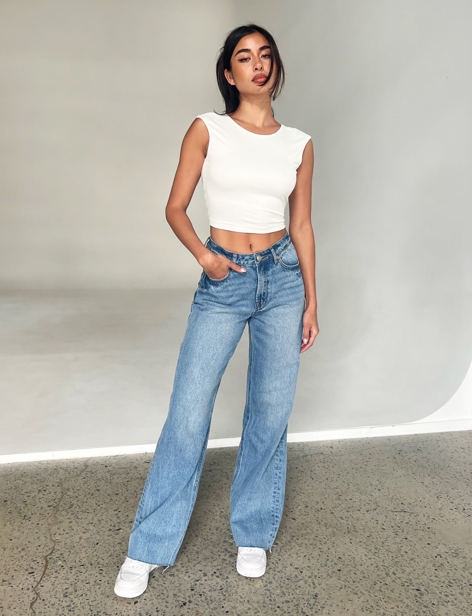 In a minimalist setting, an individual models the TIGERMIST "SACHA JEAN - MID BLUE," featuring high-waisted, relaxed straight legs with raw hemlines. They pair these jeans with a white sleeveless crop top and white sneakers. With their left hand casually in their pocket and a slight smile, they stand on a concrete floor against a plain background.