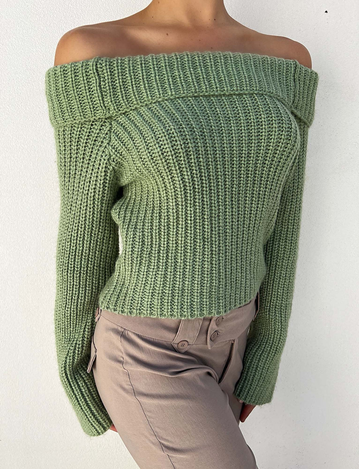 A person is wearing the ASHLYN JUMPER - GREEN from TIGERMIST, an off-the-shoulder, ribbed-knit sage green sweater with long sleeves and a thick folded neckline, paired with light brown pants. The background is plain white.