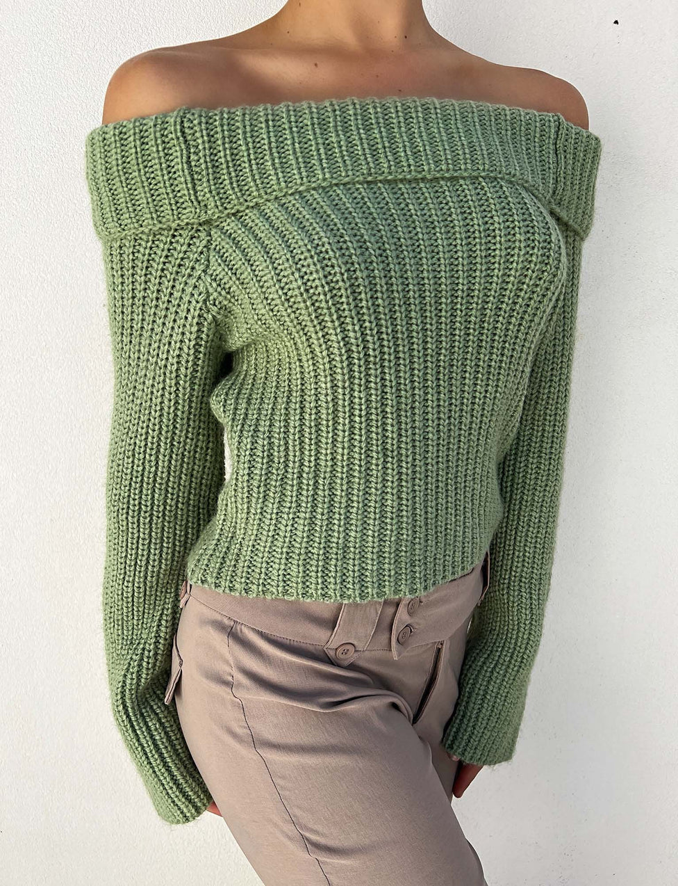 A person is wearing the ASHLYN JUMPER - GREEN from TIGERMIST, an off-the-shoulder, ribbed-knit sage green sweater with long sleeves and a thick folded neckline, paired with light brown pants. The background is plain white.