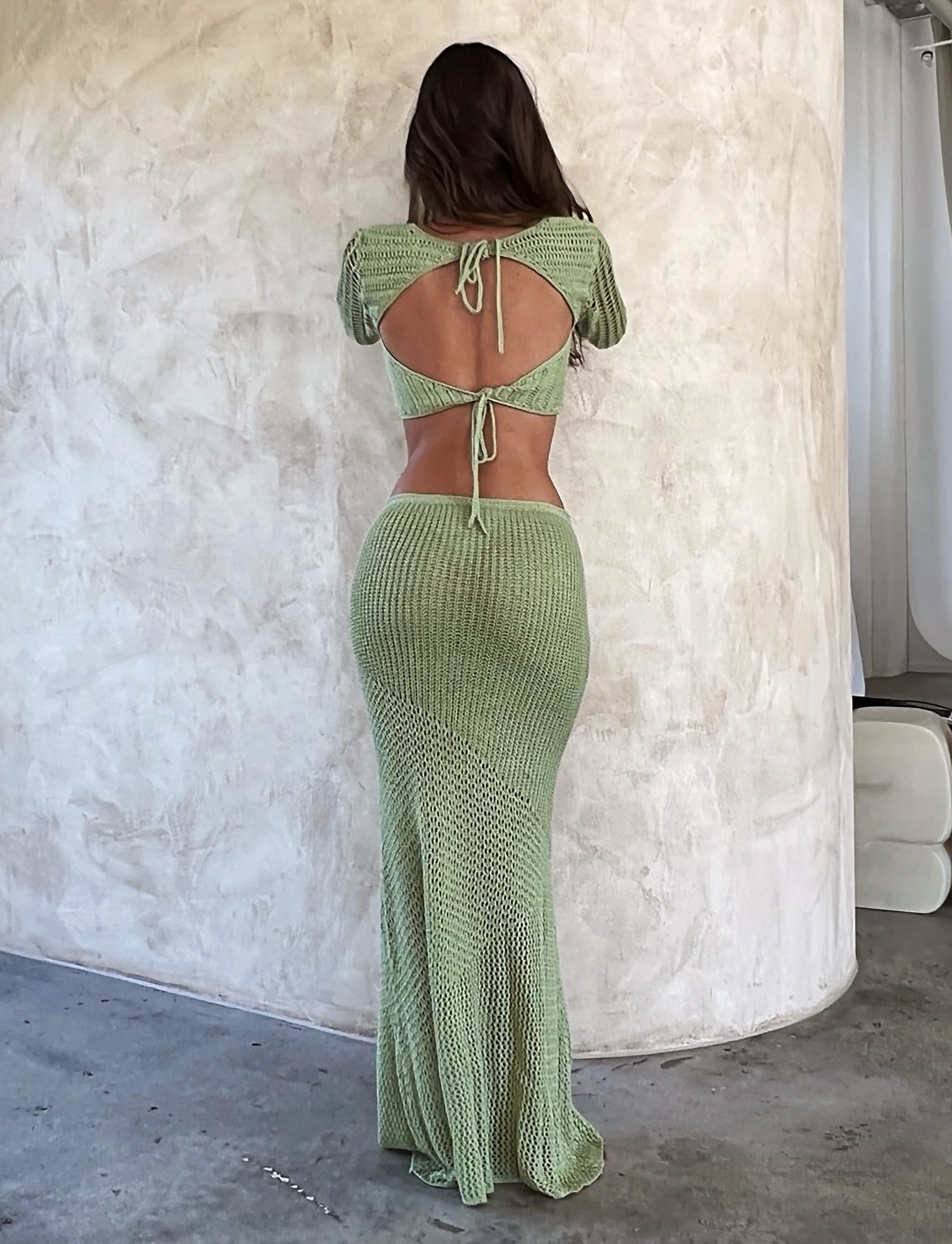 A woman with long brown hair stands with her back to the camera, posing against a textured, off-white wall. She is wearing a pastel green MACAULEY SKIRT by TIGERMIST that ties at the upper back and waist, extending down to her ankles. The setting appears to be indoors.