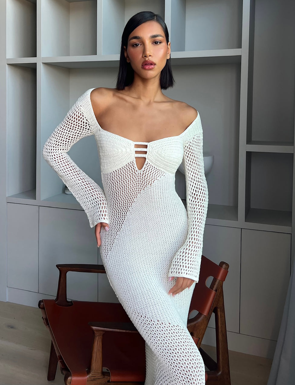 A person with straight, dark hair stands confidently by a wooden chair. They are wearing the form-fitting MACAULEY DRESS - WHITE : CREAM from TIGERMIST, featuring long sleeves and a keyhole cutout at the chest. The background showcases open cube shelving.
