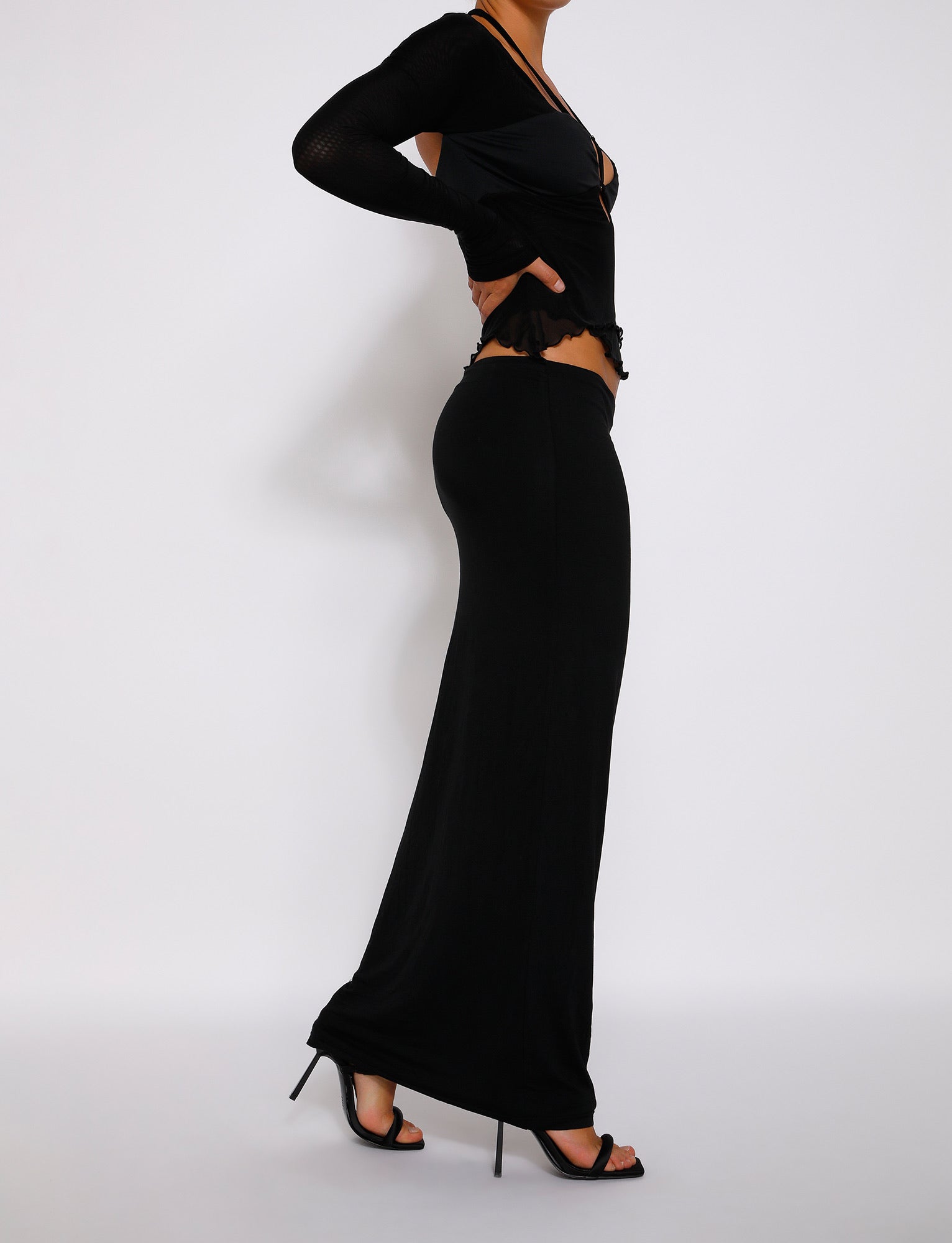 A person standing sideways against a plain white background, wearing a form-fitting black long-sleeve top and the TIGERMIST ADDISON SKIRT - BLACK with black high heels. The outfit, showcasing Tiger Mist's signature style, highlights the individual's elegant and poised demeanor.