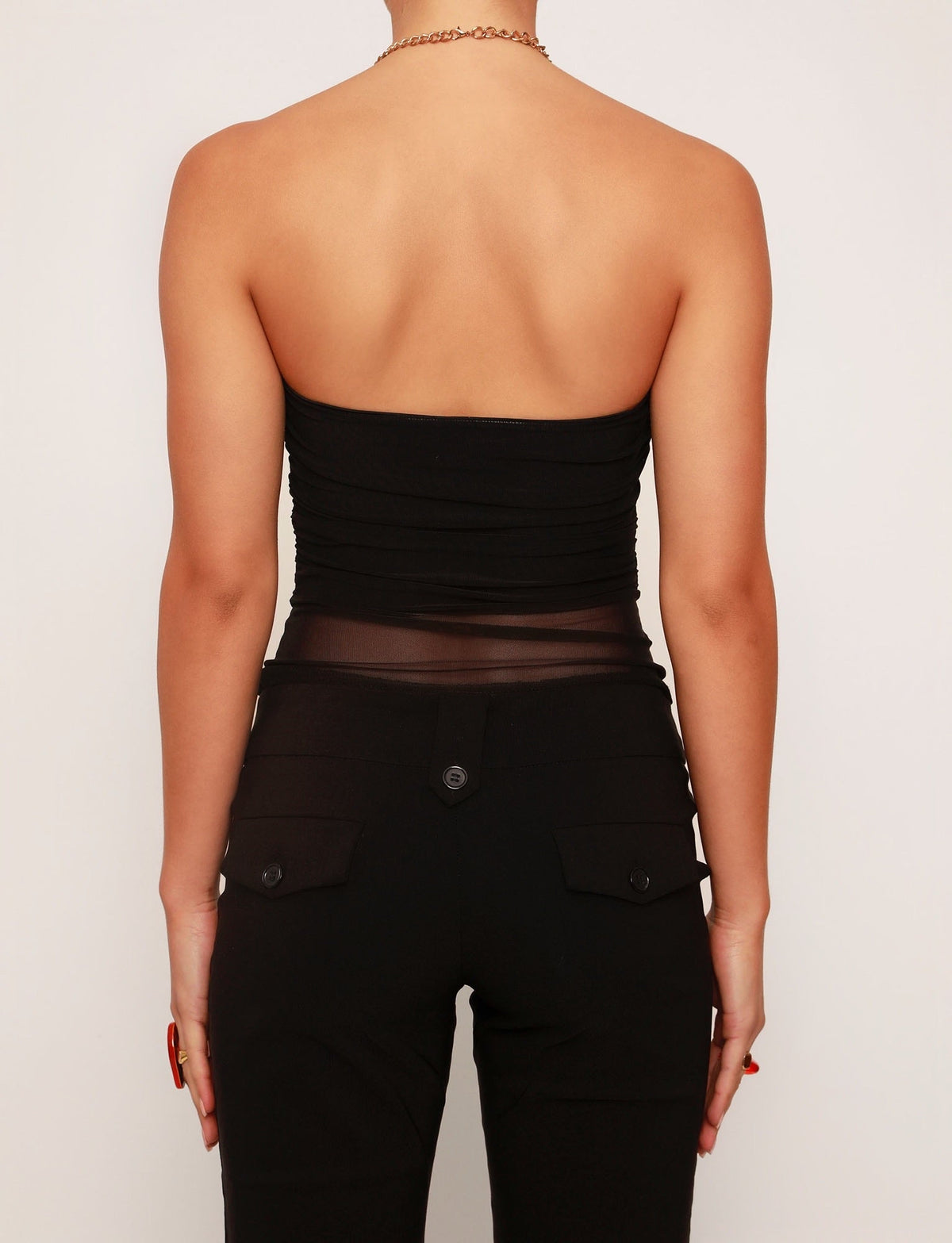 A person with short hair is seen from behind wearing the TIGERMIST HARRIETT TUBE TOP in black, paired with black pants featuring buttoned back pockets. The look is accessorized with a gold necklace and rings against a plain white background.