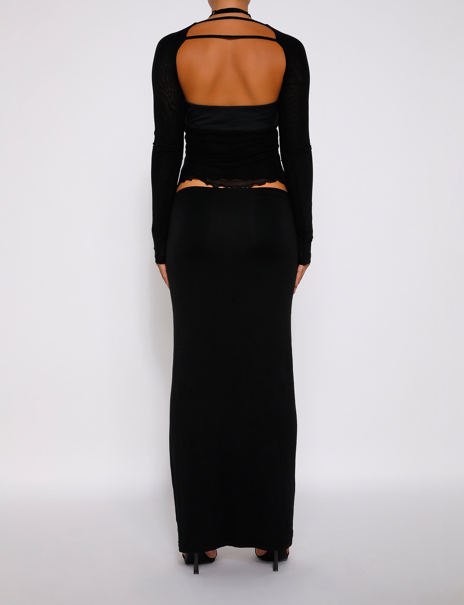 Back view of a person wearing the long-sleeve top with an open back paired with TigerMist's ADDISON SKIRT in black. They are standing against a plain white background, showcasing the clothing details and design. The person is wearing black high-heeled shoes.
