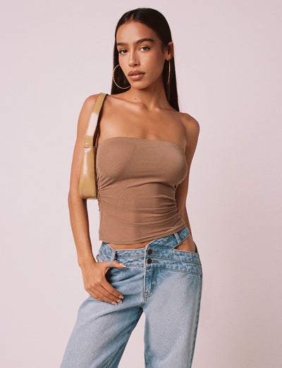 A woman with long dark hair is wearing the HARRIETT TUBE TOP in tan from TIGERMIST, paired with light blue jeans and large hoop earrings. She strikes a pose with one hand in her pocket and the other holding a beige shoulder bag strap, set against a plain, light-colored background.