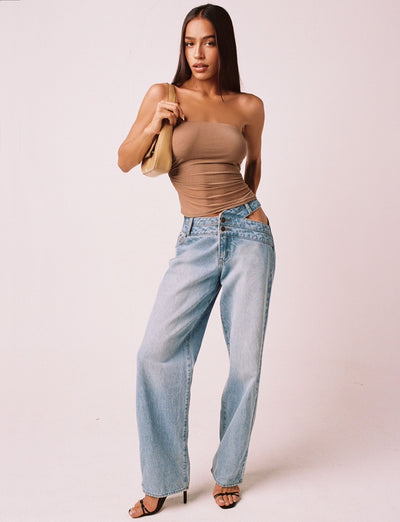 A woman with long brown hair wears a strapless TAN HARRIETT TUBE TOP from TIGERMIST and light blue, loose-fitting jeans with a cutout at the hip. She accessorizes with black strappy heels and a tan shoulder bag. She stands against a plain white background looking directly at the camera.