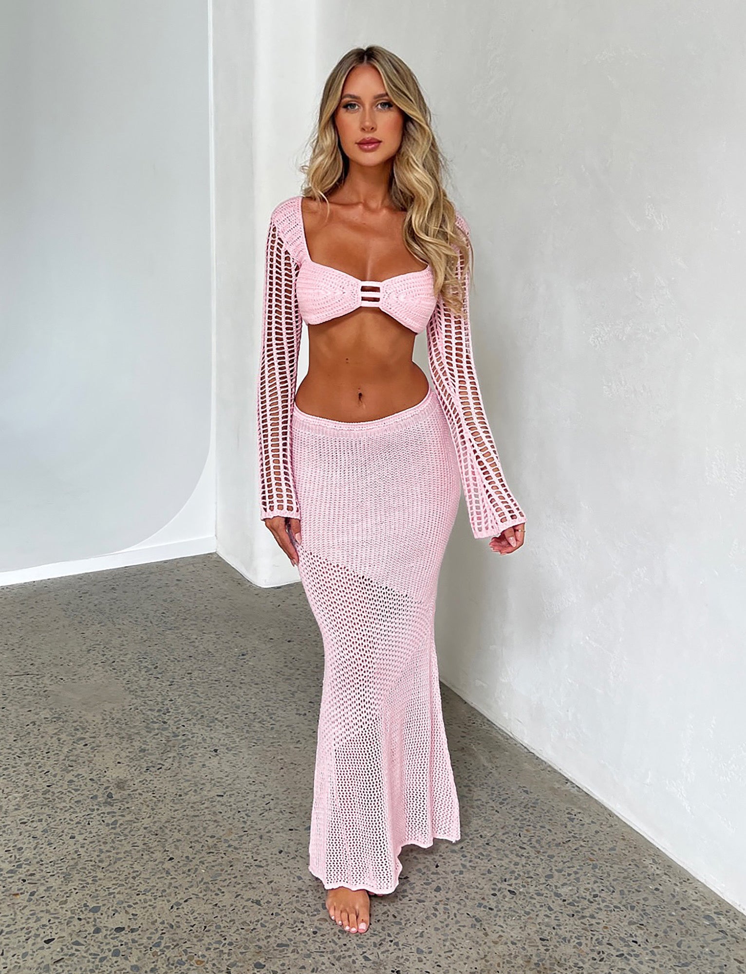A woman with long blonde hair stands against a white textured wall, wearing a pink knit bikini top with long sleeves from Tiger Mist. She pairs it with the MACAULEY SKIRT - BABY PINK, a high-waisted, floor-length skirt. The outfit features intricate knit patterns, and she stands barefoot on a speckled floor.