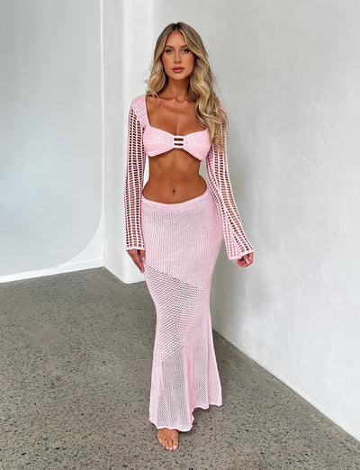 A woman with long blonde hair stands against a white textured wall, wearing a pink knit bikini top with long sleeves from Tiger Mist. She pairs it with the MACAULEY SKIRT - BABY PINK, a high-waisted, floor-length skirt. The outfit features intricate knit patterns, and she stands barefoot on a speckled floor.