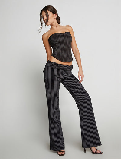 A woman stands against a plain background wearing the KITTIE PANT - PINSTRIPE by TIGERMIST, along with a matching strapless pinstripe corset top. The pinstripe design enhances her style as she looks down, one hand on her hip and the other holding the waist of her flared-leg pant.