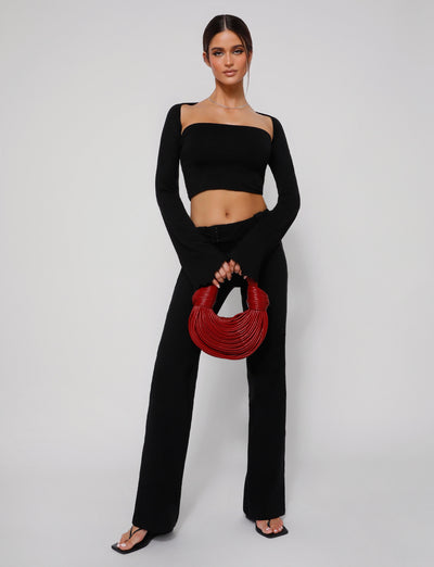 A woman stands against a plain background, wearing the SOPH TWO PIECE TOP - BLACK from TIGERMIST paired with black pants. She holds a red, circular woven handbag in front of her. With long, dark hair pulled back in a sleek style and black sandals on her feet, she exudes effortless chic.