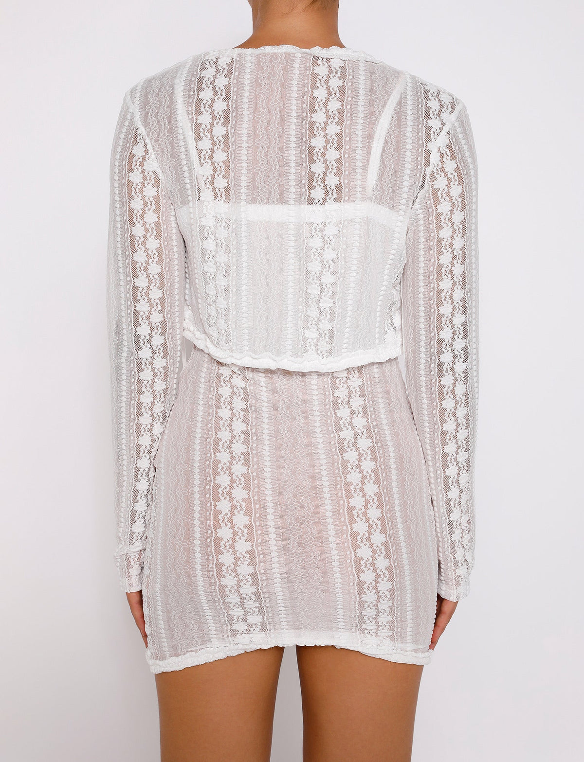 A person with tanned skin is wearing the SOPHA TWO PIECE DRESS - WHITE from TIGERMIST, a sheer white lace long-sleeved outfit featuring intricate vertical patterns. The back view reveals a boxy overlay around the shoulder area of the dress. The background is plain white.