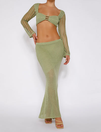A person is posing against a plain background, showcasing the MACAULEY SKIRT - GREEN from TIGERMIST, featuring a pastel green long-sleeve, open-knit crop top and matching high-waisted, open-knit floor-length skirt. The beige high-heeled sandals complement the outfit, which reveals their midriff and arms with a sheer, mesh-like texture.