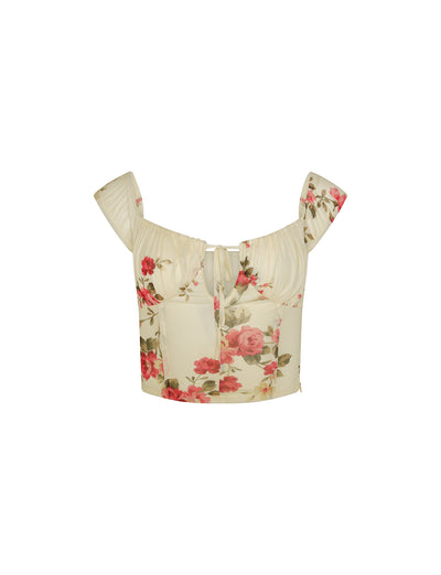 A yellow floral cropped top from TIGERMIST with pink and green rose patterns. The FEYRA TOP features short sleeves, a sweetheart neckline with a small tie in the center, and a fitted bodice.