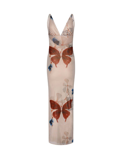 A woman stands against a textured grey wall, wearing the Percy Maxi Dress by Poppy Butterfly. The sleeveless, bodycon fit dress features a deep V-neckline and a side slit. The cream-colored maxi dress showcases a paisley print with blue and rust accents. Her blonde hair is styled in loose waves.